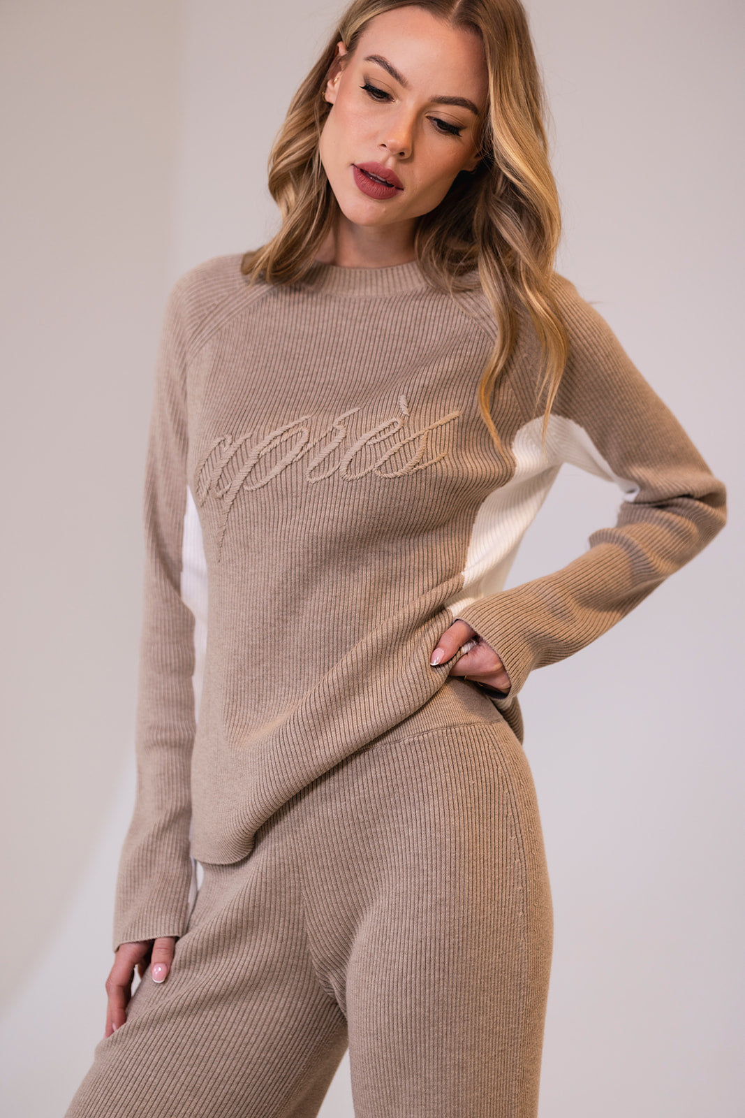 Woman wearing a beige ribbed sweater and pants with 'après' embroidered text