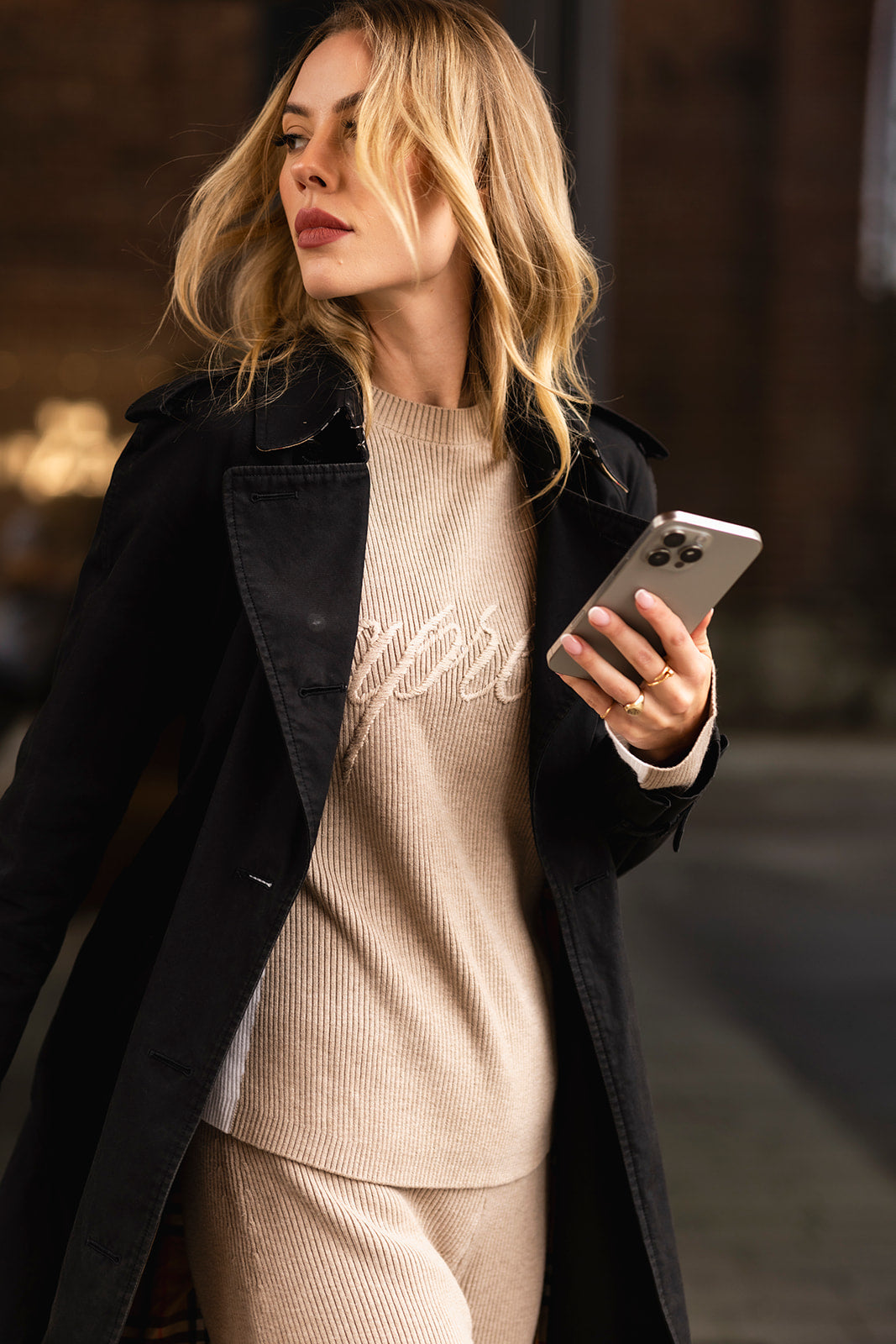 Woman holding a phone, wearing a black coat and beige ribbed knit sweater with &#39;Apres&#39; embroidered branding.